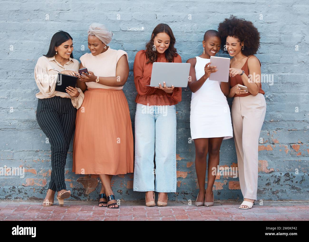 Group of five young happy cheerful businesswomen standing against a wall outside in the city and using tech. Happy businesspeople talking and using Stock Photo