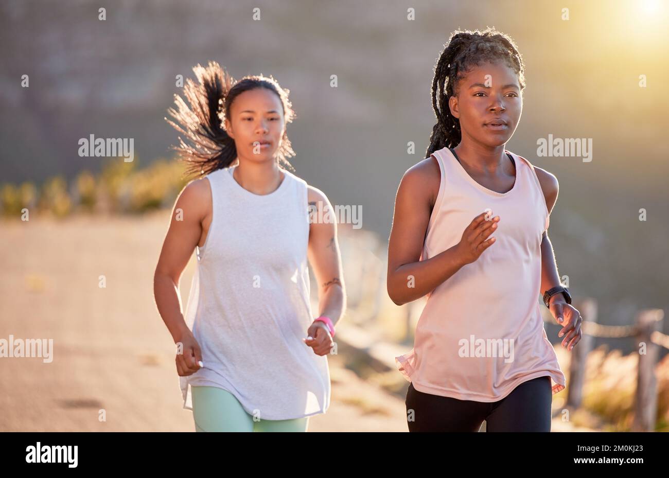 Two focused young female athletes out for a run on a mountain road on a sunny day. Energetic young women running outdoors to help their bodies in Stock Photo