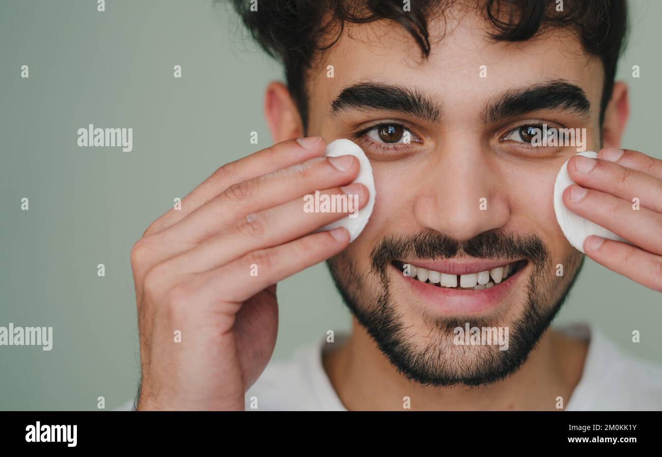 Portrait of a smiling man cleaning his face with two cotton pads on gray background. Skin care healthcare cosmetic procedures concept Stock Photo