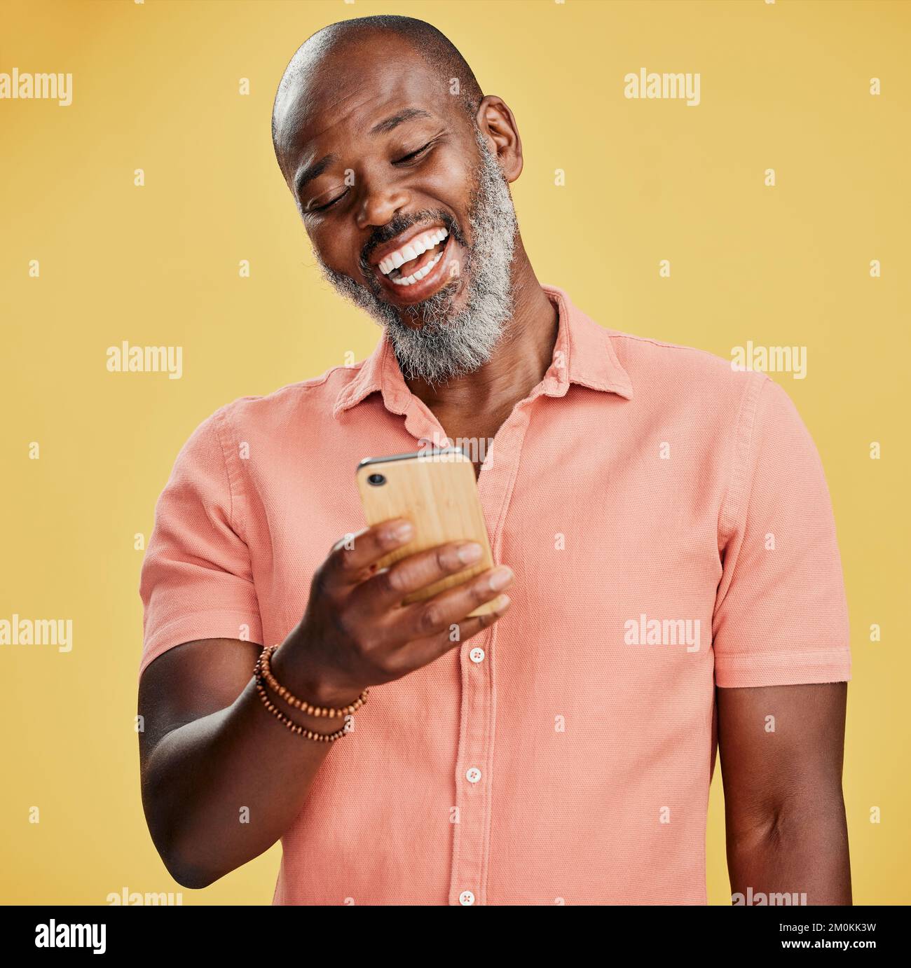 One happy African American man standing against a yellow studio background, holding and using his cellphone to browse the internet. Smiling black man Stock Photo