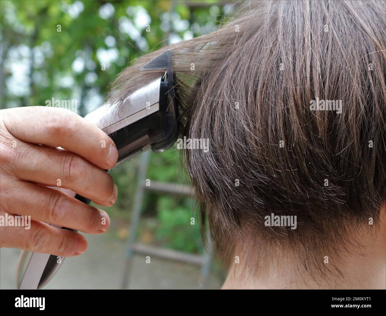 a man's hand holds a trimmer and cuts long brown hair on the back of a man's head, cutting long hair on a teenager's head on the street Stock Photo