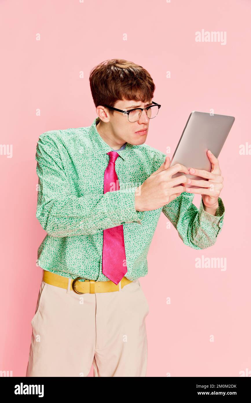 Funny emotional man, student geek wearing vintage style shirt and trousers having fun, using tablet isolated over light pink background. Fun, style Stock Photo