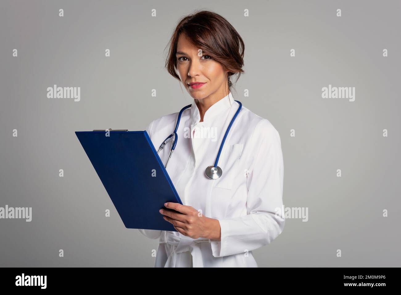 Studio portrait of smiling female doctor holdig clipboard in her hand and wearing labcoat while standing at isolated grey background. Copy space. Stock Photo