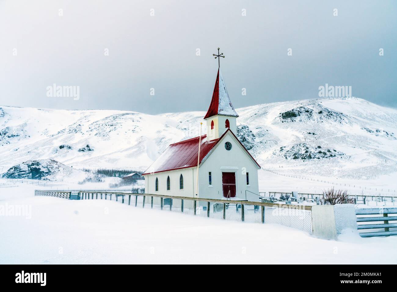 Small church with bright red roof stands in contrast to snow covered field and mountains in a small village in Iceland. Stock Photo