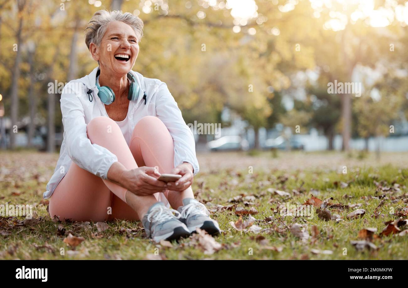 Health, nature and senior woman in park with phone and headphones to relax during fitness workout. Music, happiness and old lady laughing, sitting on Stock Photo