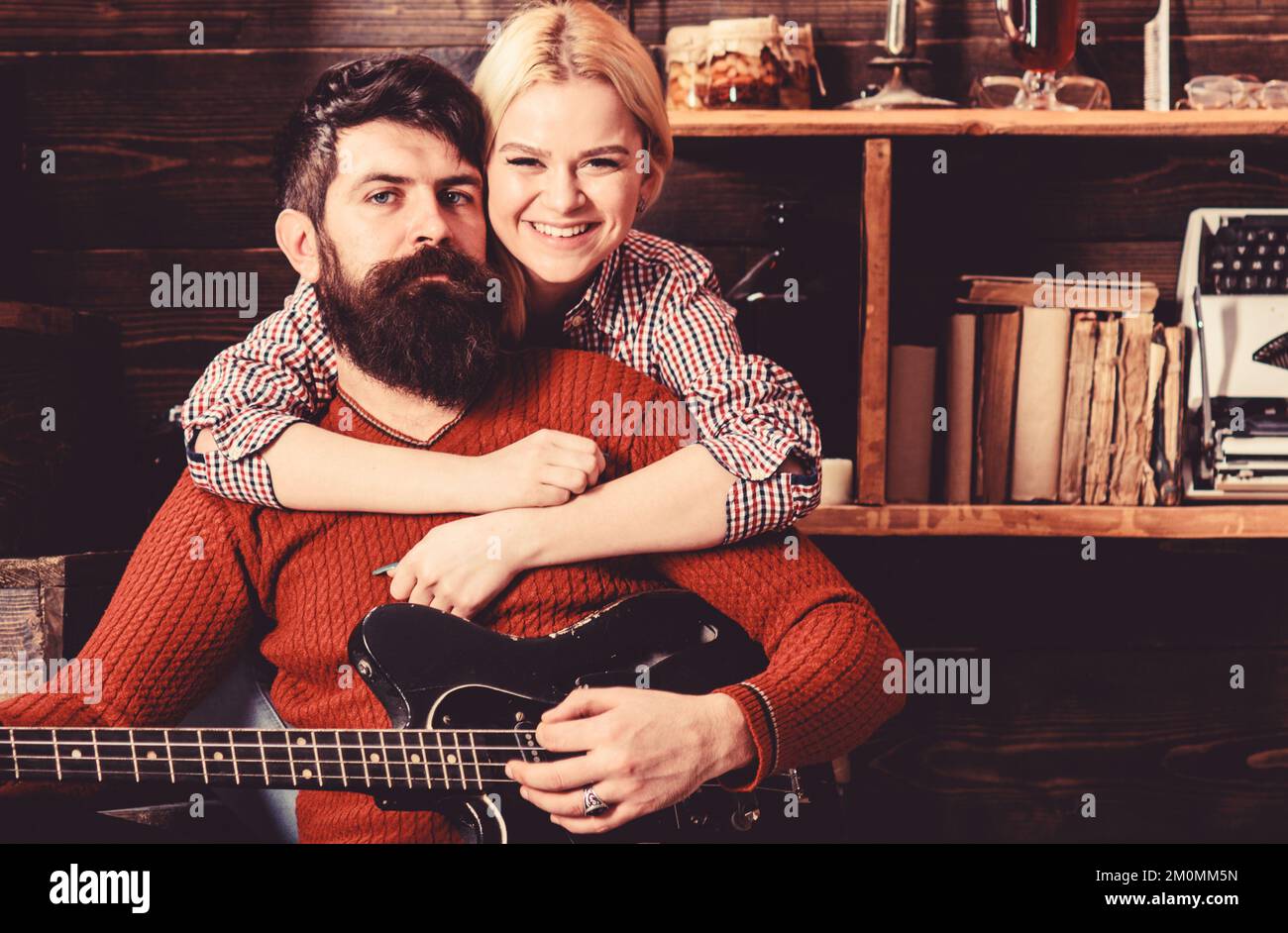 Romantic evening concept. Lady and man with beard on happy faces hugs and plays guitar. Couple in wooden vintage interior enjoy guitar music. Couple Stock Photo