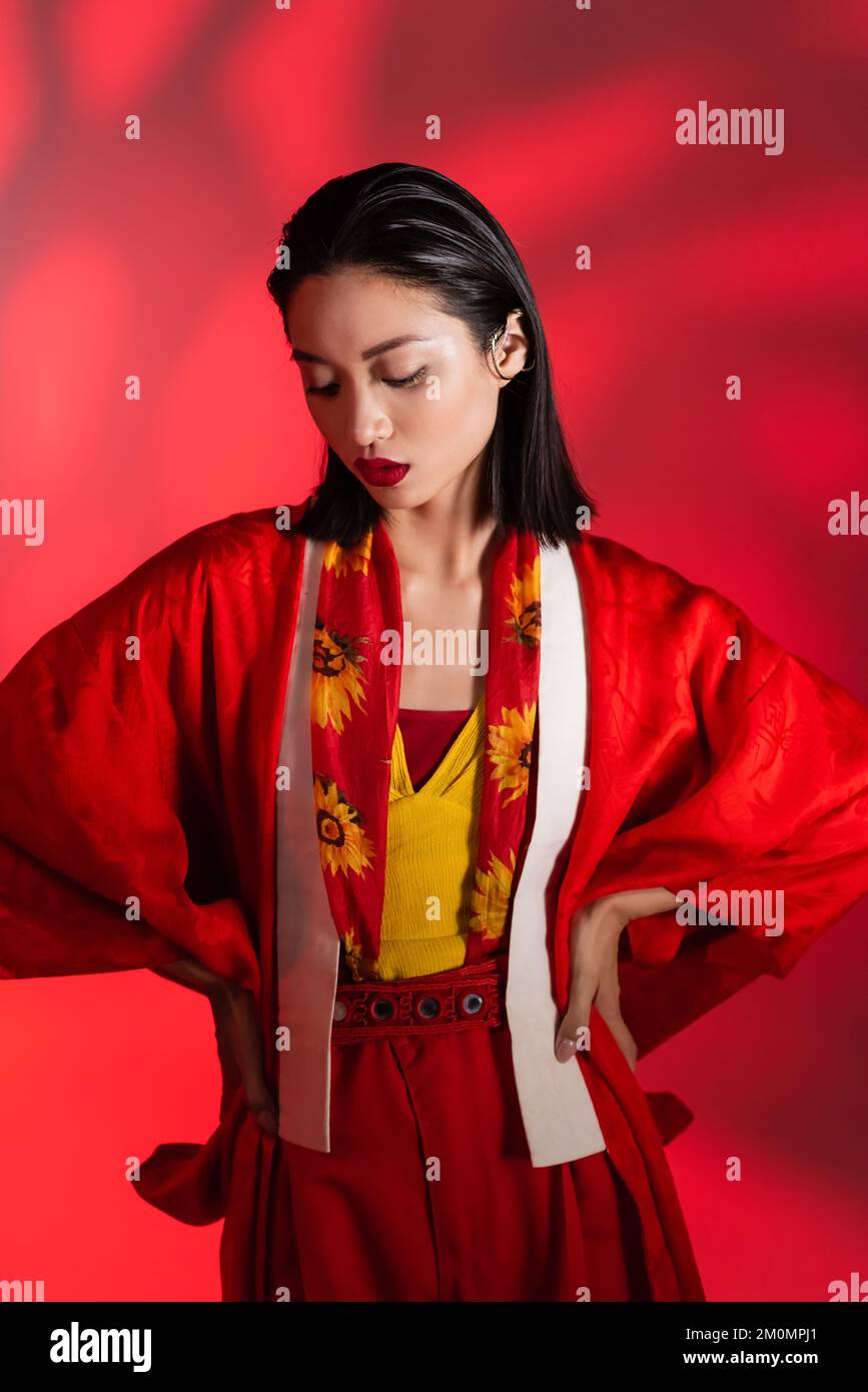 brunette asian woman in neckerchief with floral decor and kimono cape posing with hands on hips on abstract red background,stock image Stock Photo
