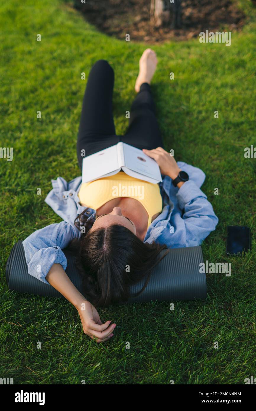 Top view of female in sporty clothes lying on green grass reading book, she take a deep breath on fresh air. Beautiful green. Fitness woman. Nature Stock Photo
