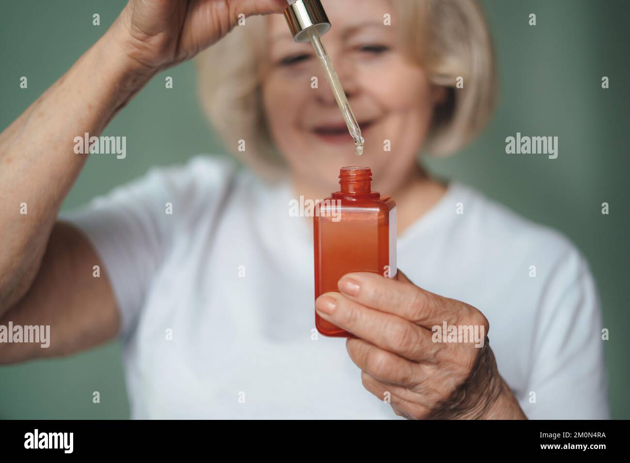 Close-up portrait older female's hands holding red bottle with facial essence dropping collagen moisturizer face serum. Rejuvenation and skincare Stock Photo