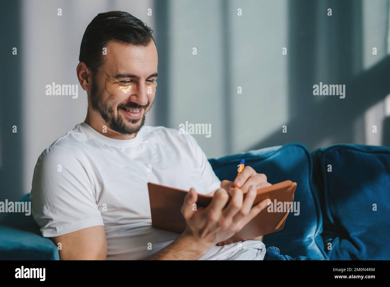 Smiling young student man wearing white t-shirt sitting on blue sofa indoors writing down memories in notebook diary, resting on weekends at home Stock Photo