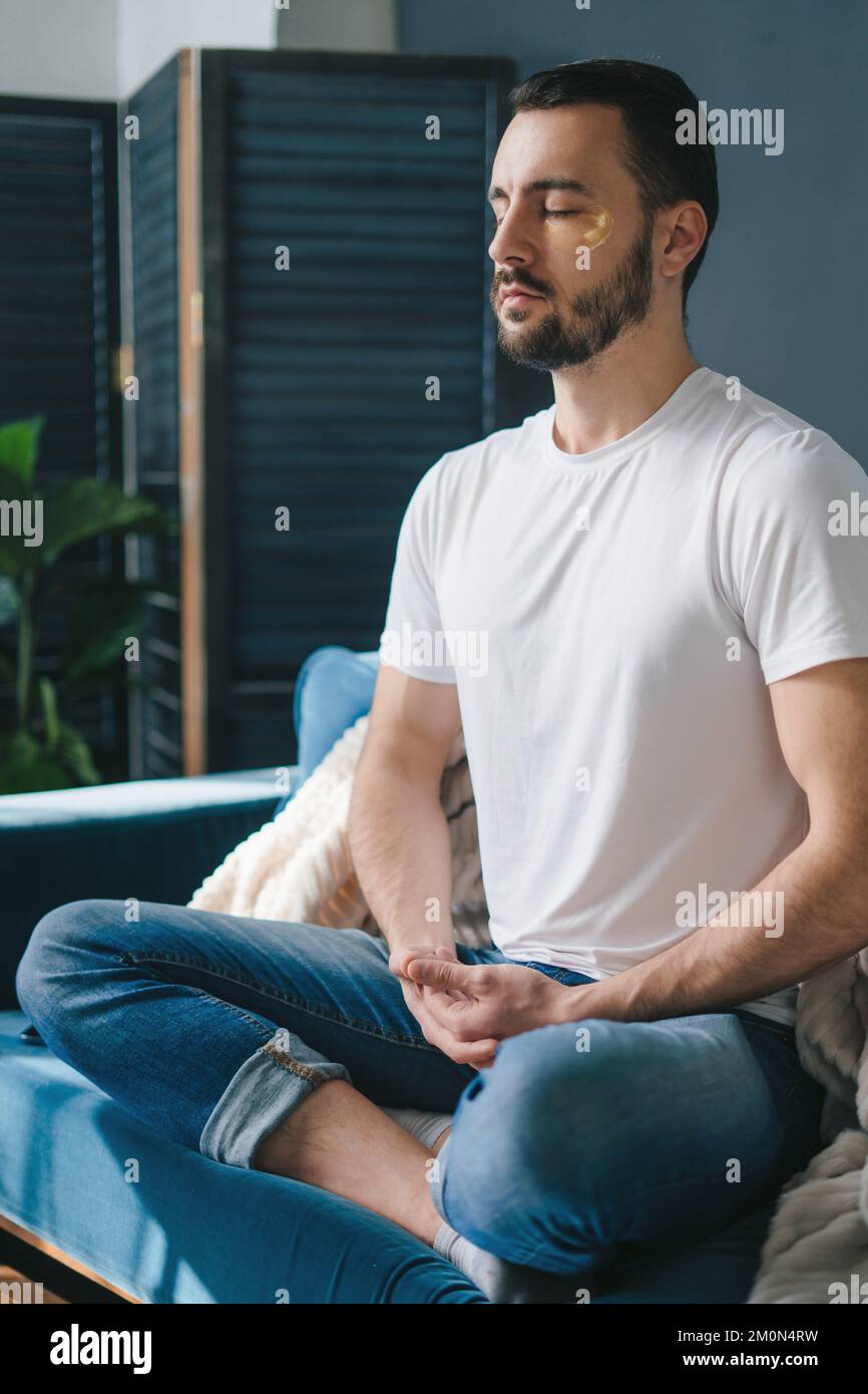 Handsome man practicing meditation in the cross-legged posture, on the sofa in his living room. Breathing exercise. Stock Photo