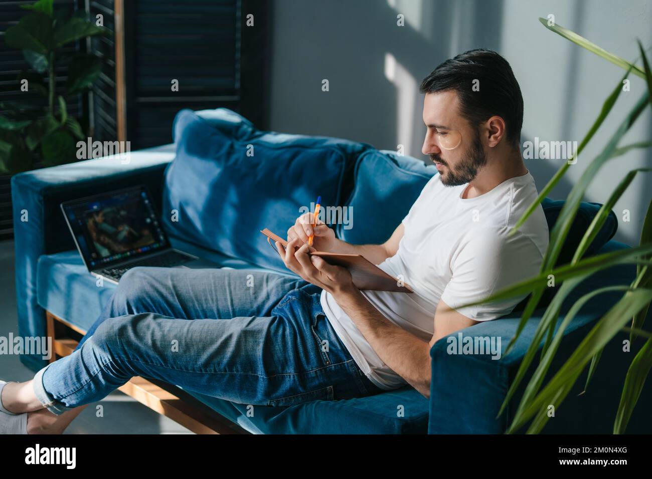 Young man sitting on sofa writing down information in red note book, taking important notes with pencil. Resting on weekends staying at home. Stock Photo