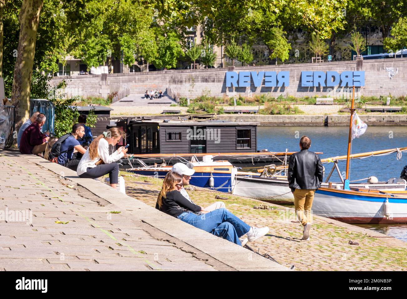 Young people enjoy the sunshine sitting on the banks of the Erdre river in Nantes, France, with the large sign 'REVER ERDRE' in the background. Stock Photo
