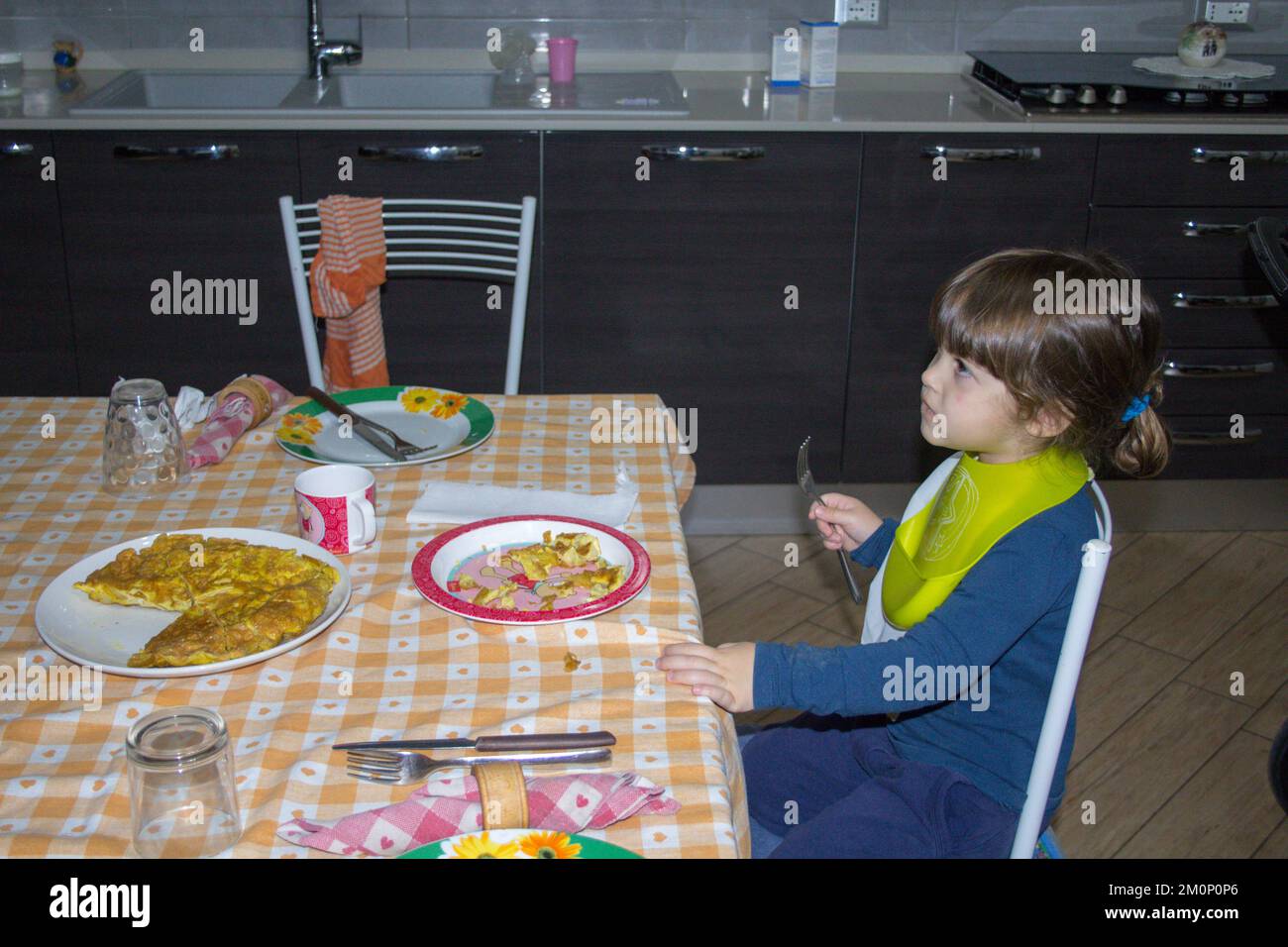 Image of an adorable little brunette girl wearing bibs, sitting at the table eating an omelette and enthralled watching tv. Distraction of children Stock Photo