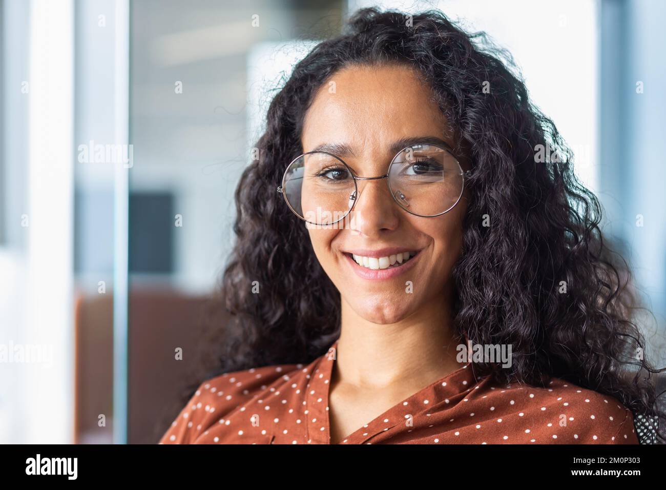 Close up photo portrait of beautiful Latin American woman with curly hair and glasses, businesswoman inside office building smiling and looking at camera. Stock Photo