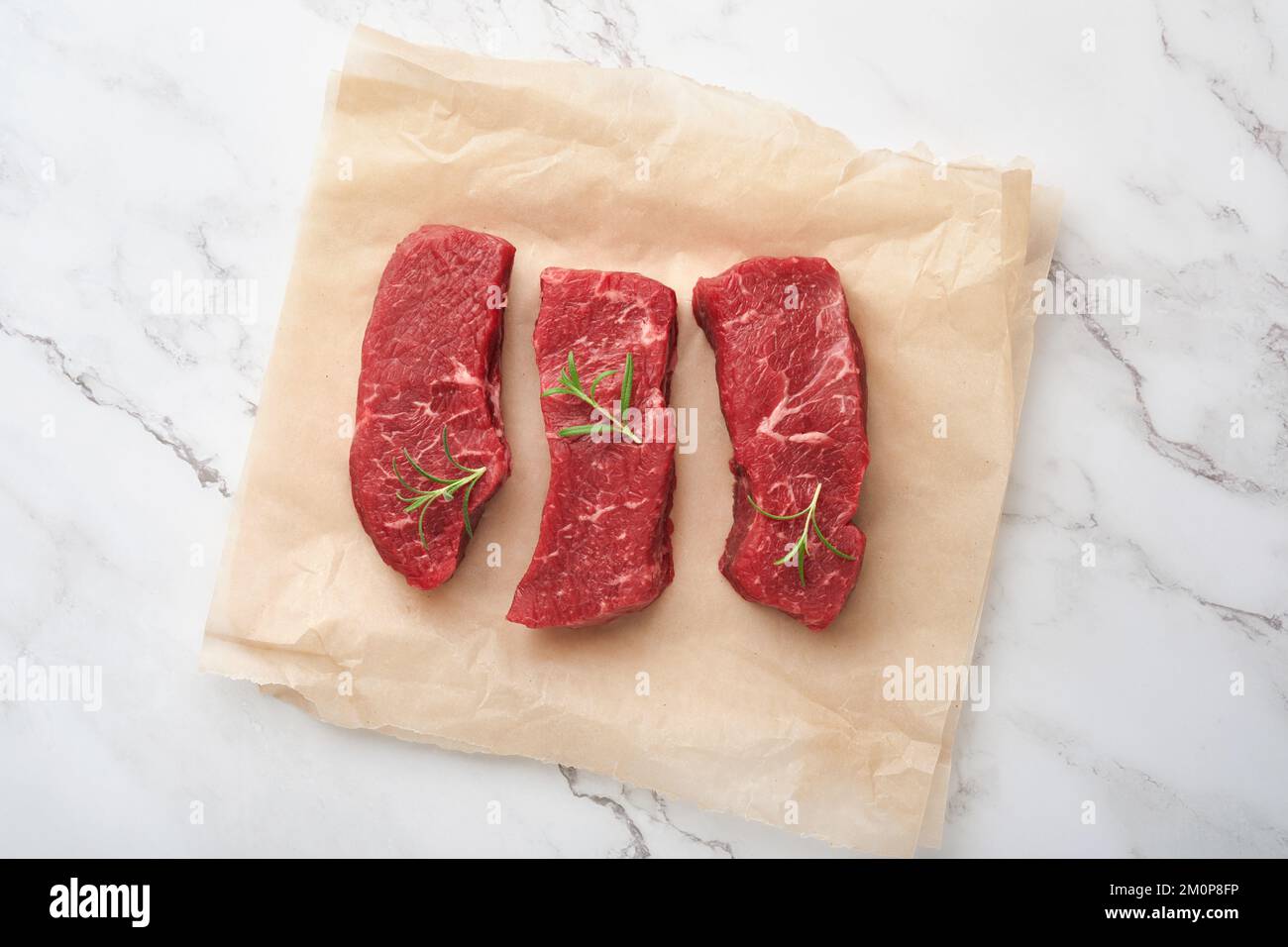Raw steaks. Top blade steaks on wood burning board with spices, rosemary, vegetables and ingredients for cooking on parchment paper on white backgroun Stock Photo