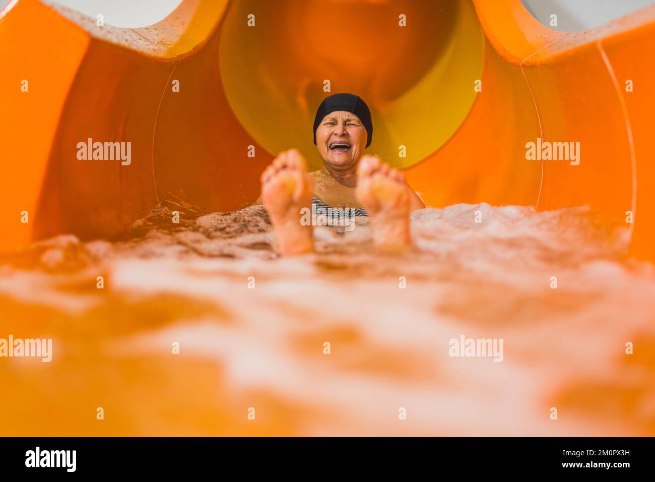 Fun swimming pool or aqua park activities. Laughing amused elderly caucasian female pensioner in black hair cap sliding on an orange water slide. Blurred feet in the foreground. High quality photo Stock Photo