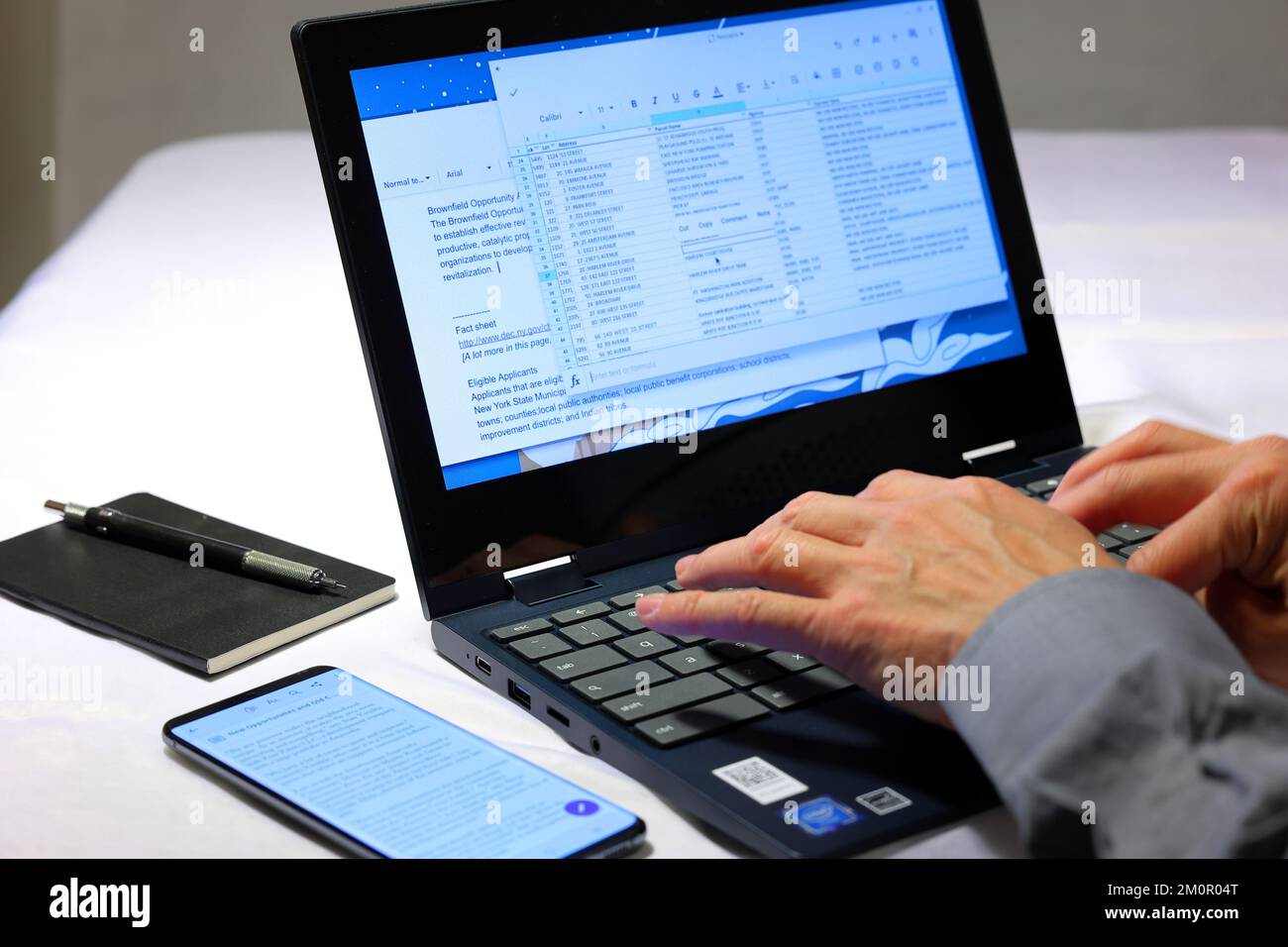 A closeup of a person's hands typing into a laptop computer with a notebook and smartphone nearby. Stock Photo