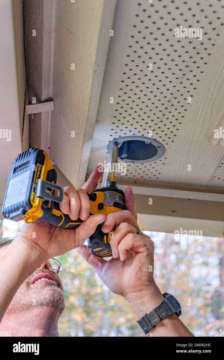 Electrician or handyman at work installing outside security lights using a battery powered electric drill or screwdriver to make the installation. Stock Photo