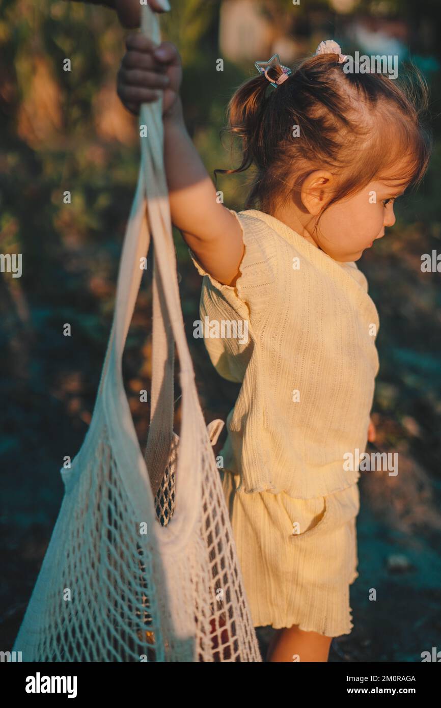 Baby girl holding a string shopping bag with vegetables in the garden at sunset. Food concept. Healthy nutrition garden food. Health concept. Stock Photo