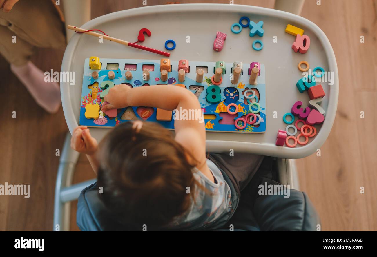 Top view of a baby girl sitting on high-chair playing with wooden cubes with numbers and colorful toy bricks. School education. Happy lifestyle Stock Photo