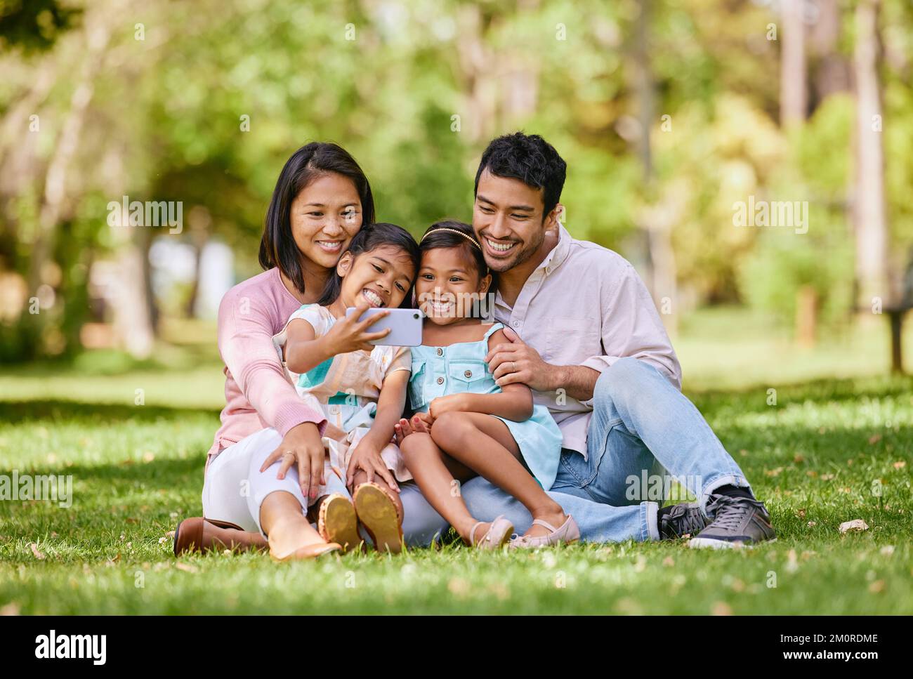 Happy asian family taking selfies on a cellphone in a park. Adorable little girls bonding with their parents outside. Full length husband and wife Stock Photo
