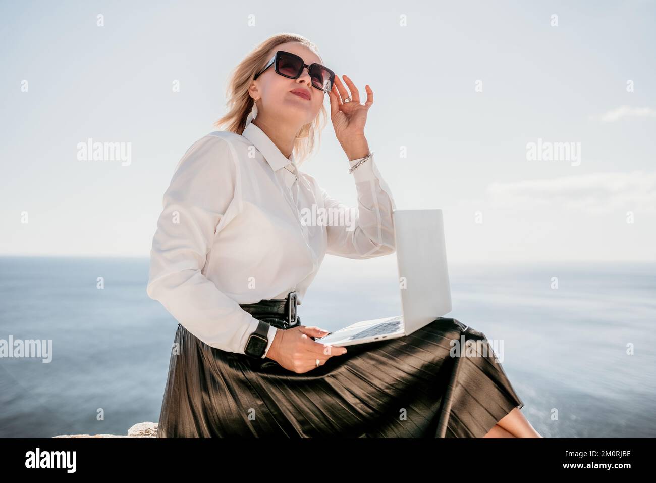 Digital nomad, Business woman working on laptop by the sea. Pretty lady typing on computer by the sea at sunset, makes a business transaction online Stock Photo