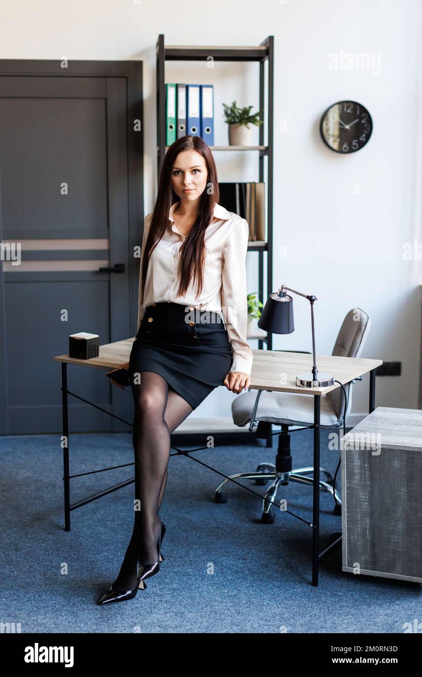 A young woman, leaning on a desk, dressed in a business style, looks confidently into the camera. Stock Photo
