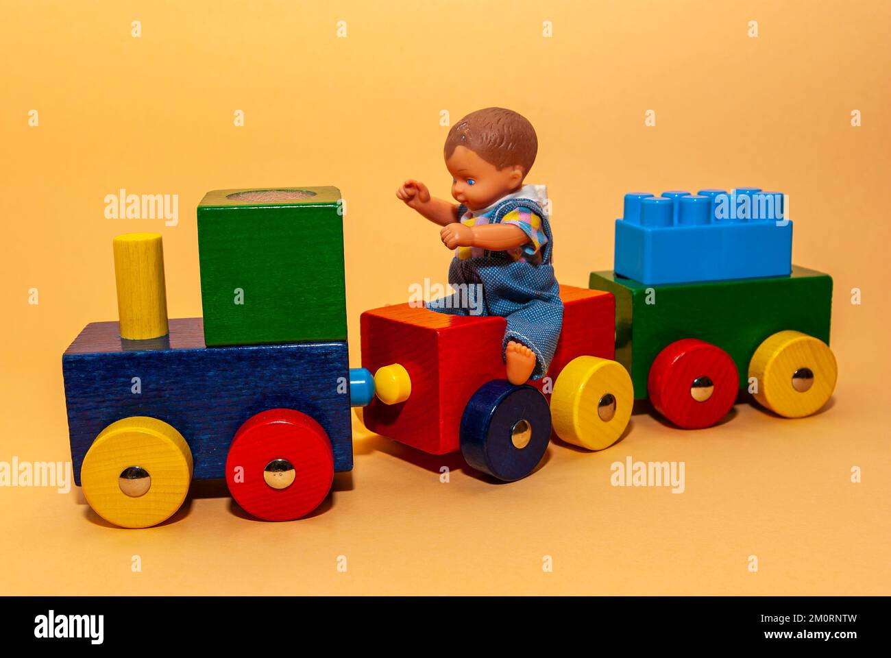 A little boy doll sits on a toy train made of wood, which has a plastic building block loaded, also concept of play and rail travel. Stock Photo