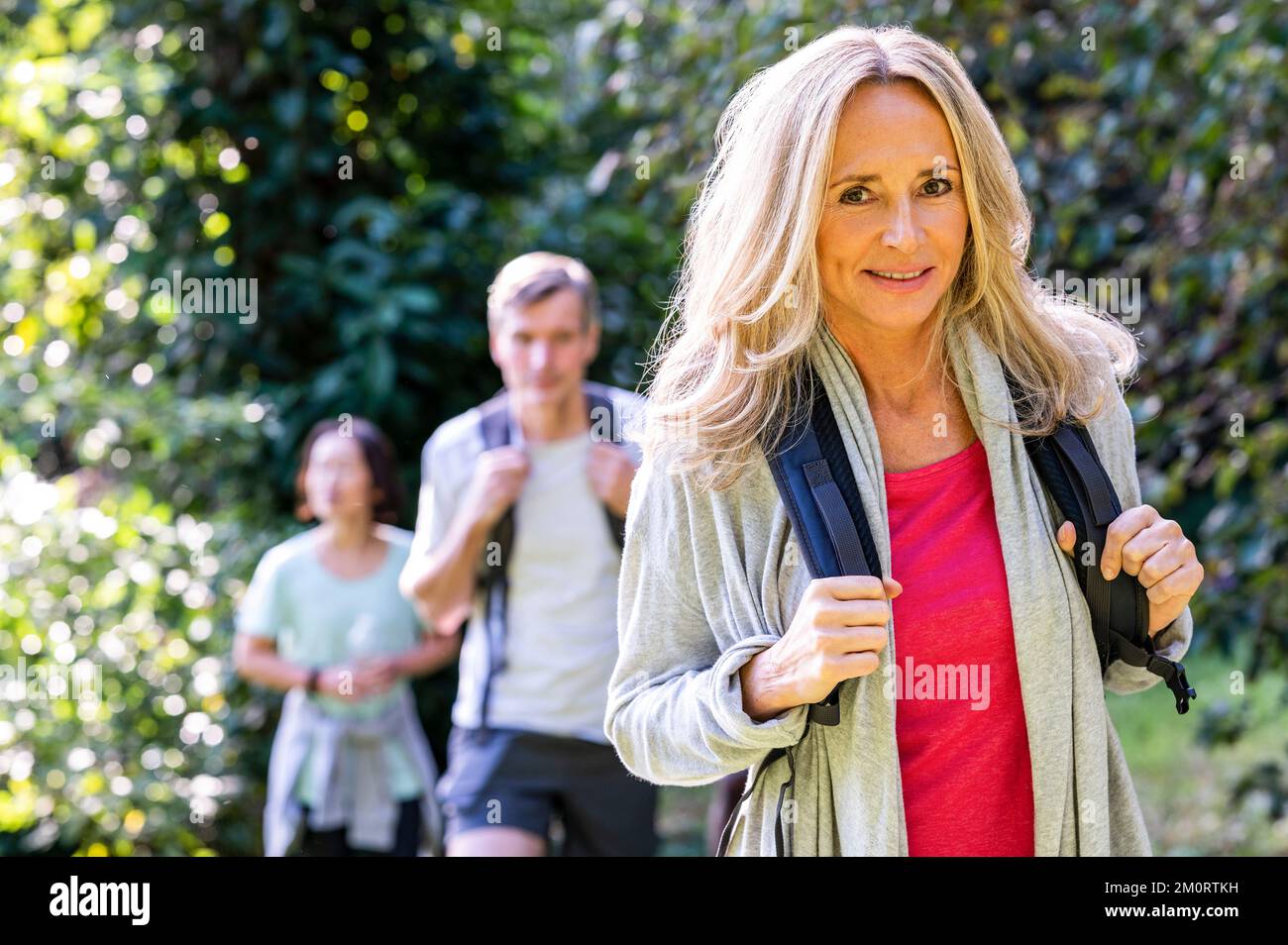 Mid-shot of blonde middle-age woman with backback hiking with her diverse group of friends in the woods Stock Photo