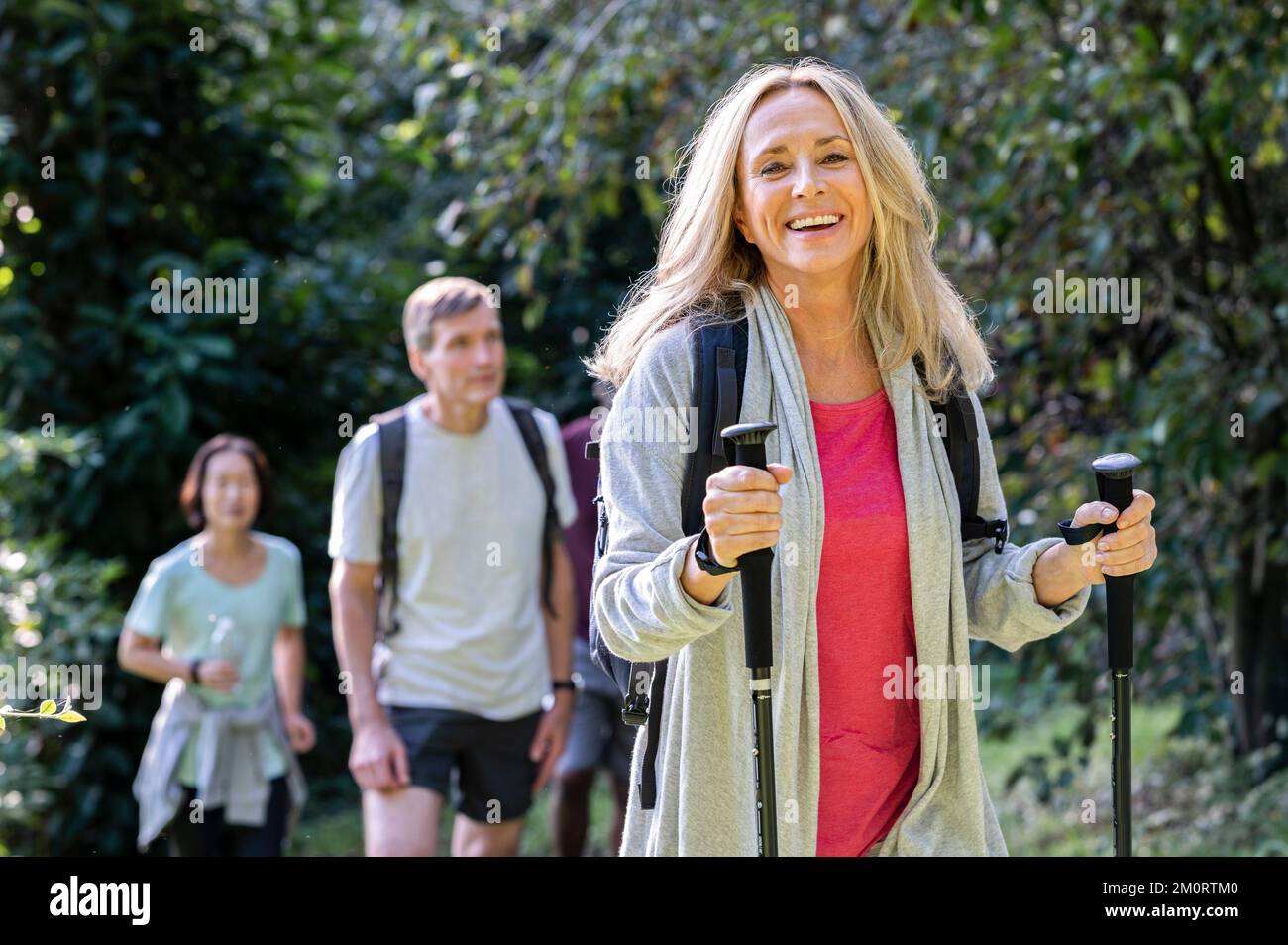 Blonde middle aged lady carrying backback and hiking poles hiking in the woods with group of friends Stock Photo