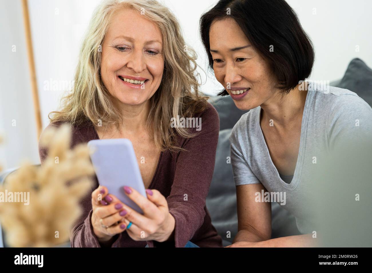 Portrait of two middle age female friends looking a mobile phone screen inside living room Stock Photo