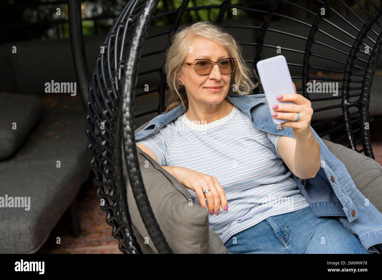 Senior woman sitting in comfortable garden chair while taking a selfie Stock Photo