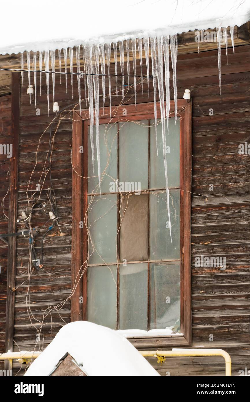 Small icicles hang on the edge of the roof, winter or spring. Log wall of an old wooden house with windows. Large cascades of icicles in smooth, beautiful rows. Cloudy winter day, soft light. Stock Photo