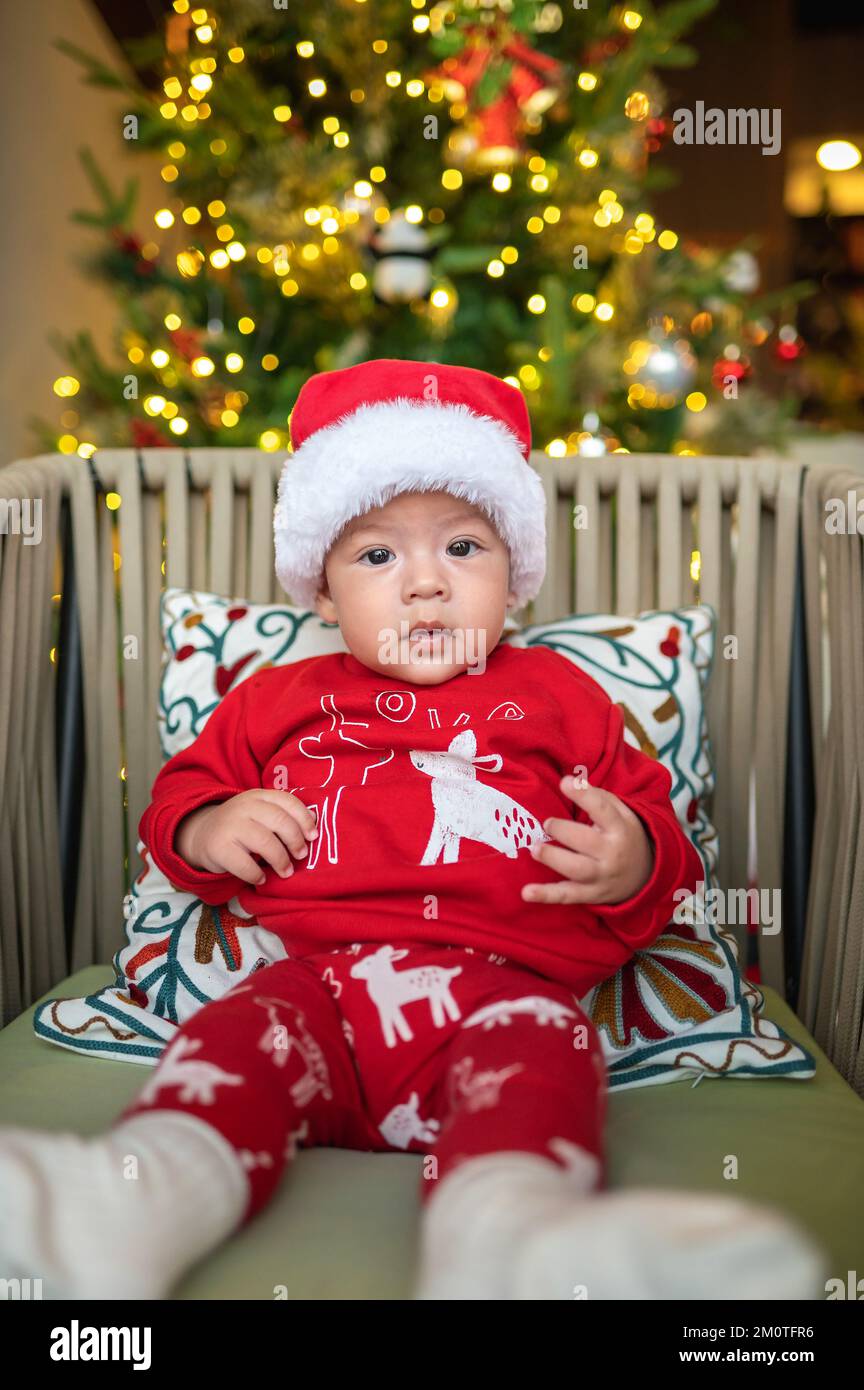 Cute newborn baby boy wearing Santa hat and festive outfit next to a Christmas tree with festive decoration and ferry lights for New Year and Christma Stock Photo