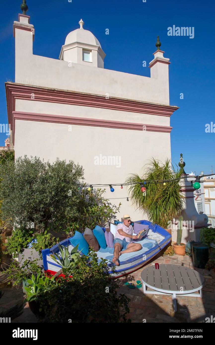 Spain, Andalusia, Cadiz, casa del consul, man in shorts and straw hat with a book in his hand lying in a boat on the tree-lined terrace of the boutique hotel la casa del consul at the foot of an 18th century shopping tower Stock Photo