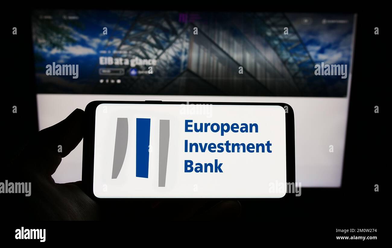 Person holding smartphone with logo of EU institution European Investment Bank (EIB) on screen in front of website. Focus on phone display. Stock Photo