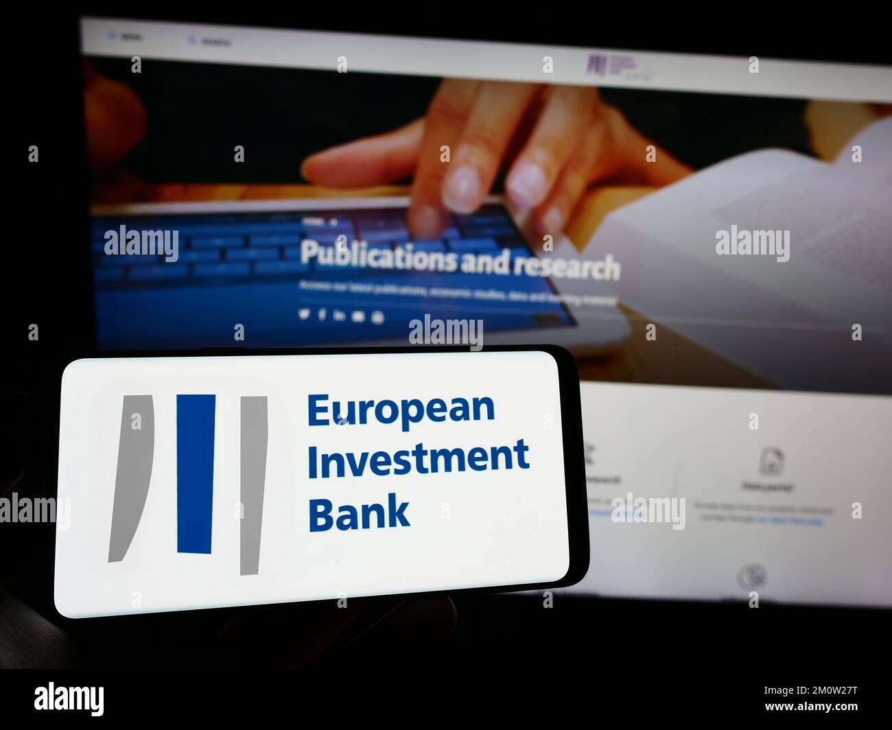 Person holding cellphone with logo of EU institution European Investment Bank (EIB) on screen in front of webpage. Focus on phone display. Stock Photo