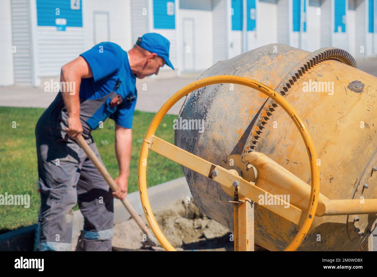 Uniformed construction worker works on construction site on summer day. Elderly bricklayer shovels cement and sand into concrete mixer. Authentic workflow. Background.. Stock Photo