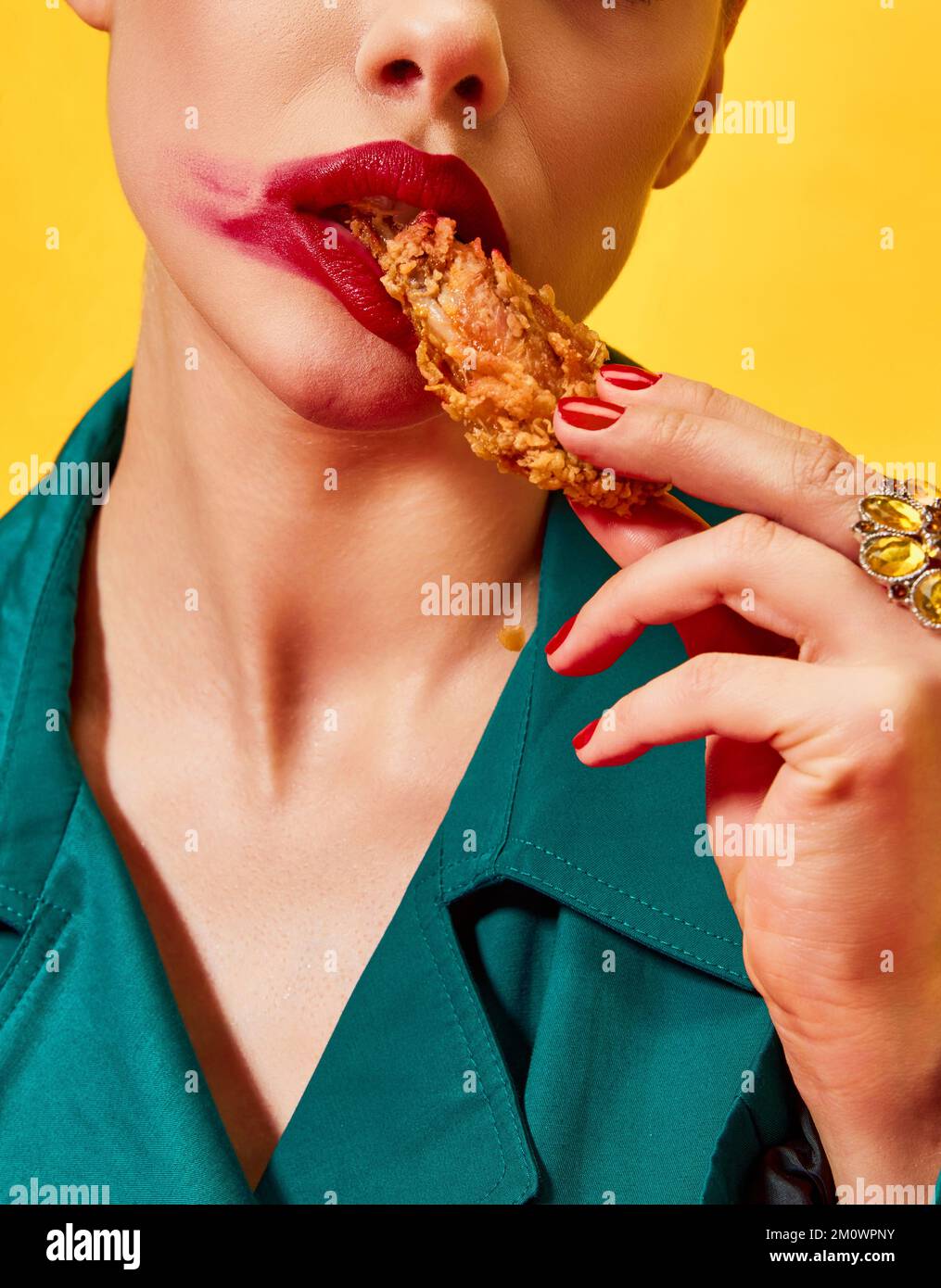 Cropped image of woman in green coat with red lipstick smudge, eating fried chicken, nuggets over yellow background. Fast food. Food pop art Stock Photo