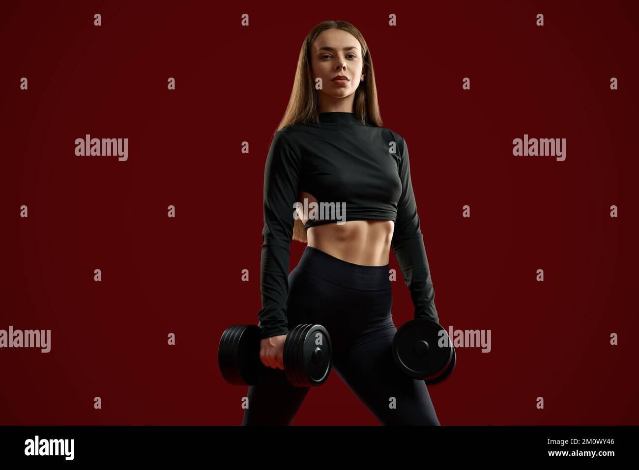 Front view of pretty brunette woman in black posing with dumbbells on red background. Beautiful sporty female with sports equipment looking seriously at camera. Concept of sport trainer lifestyle. Stock Photo