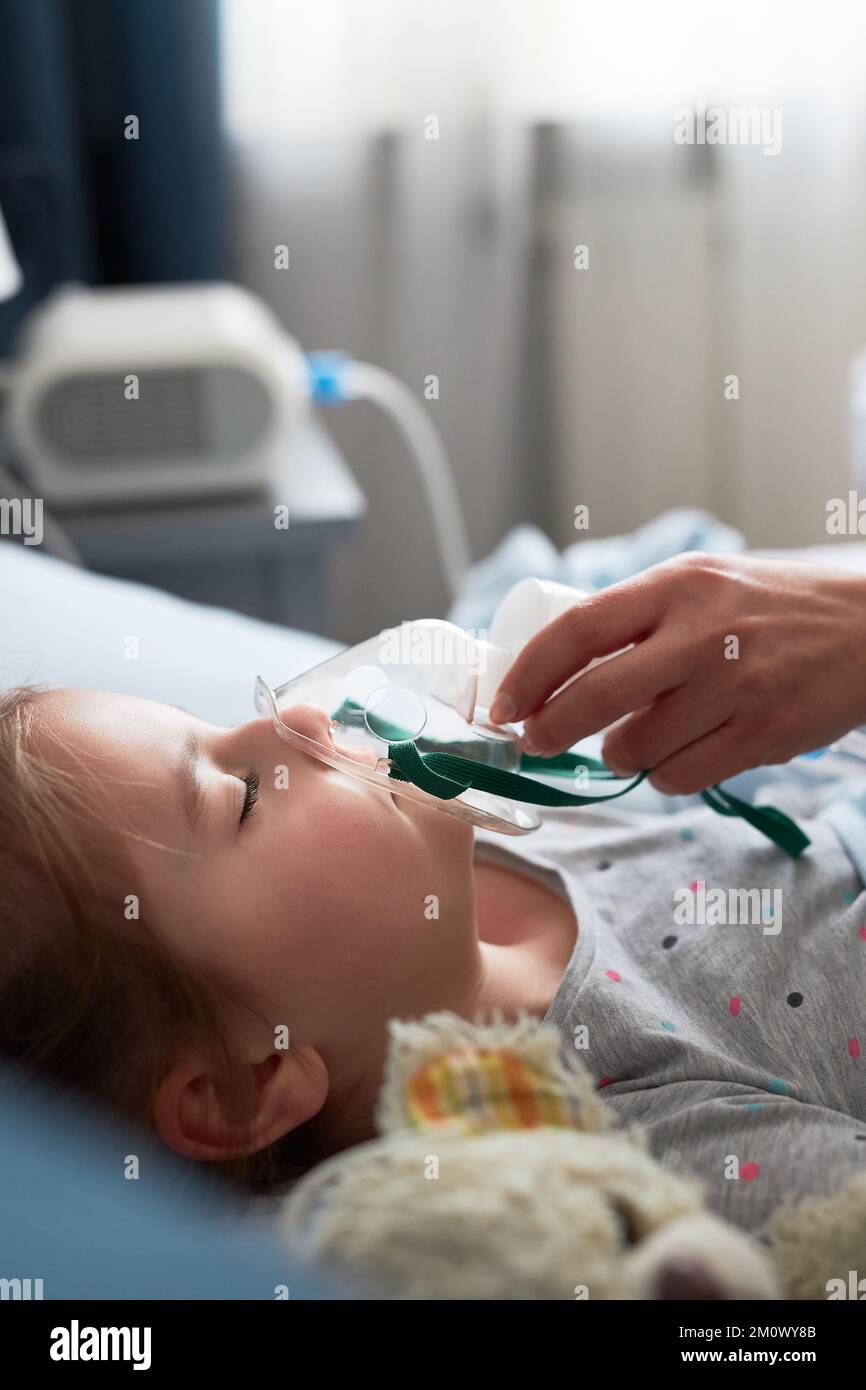 Doctor visiting little patient at home. Child having medical inhalation treatment with nebuliser. Girl with breathing mask on her face. Woman wearing Stock Photo