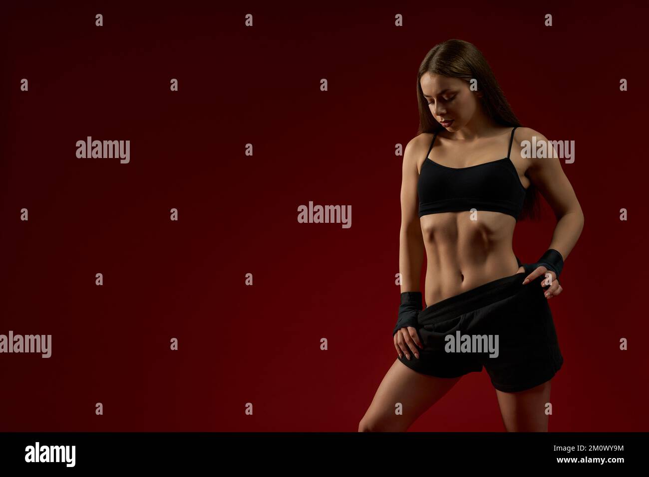 Seductive confident female trainer in black sport wear for kickboxing posing on red studio background and smiling. Sexy confident sporty woman posing looking down. Concept of box. Stock Photo