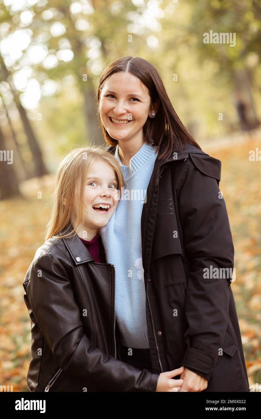 Vertical smiling cheerful glad, satisfied family of mother and small blond daughter hugging, look at camera, stand close Stock Photo