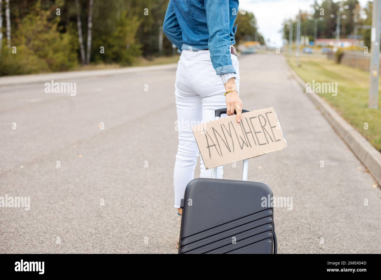Rearview cropped woman hitchhiker with plate anywhere carry roll aboard suitcase, walk by asphalt road. Adventure time Stock Photo