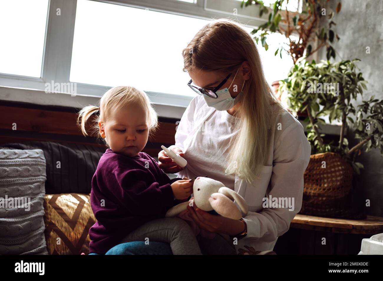 Portrait of little girl toddler sitting on childrens doctor wearing medical mask refusing to get nasal medicine at home. Stock Photo