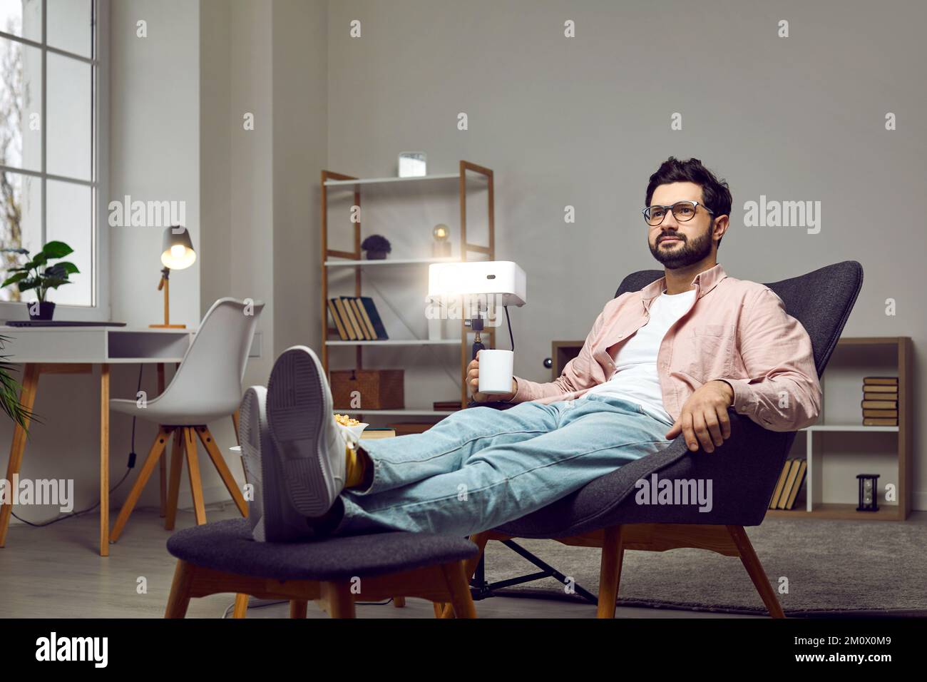 Relaxed man sitting in an armchair at home, using a projector, watching ...