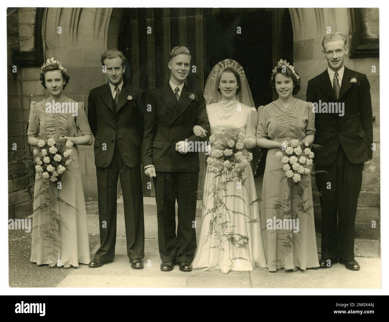 Original WW2 era wedding photograph group portrait of a happy pretty bride, wearing a white bridal gown, with puff sleeves and headdress with veil, bridegroom and best man in suits and attractive bridesmaids again wearing dresses with puff sleeves, standing outside a church, from London and Northern Studios, Alnwick, Northumberland, Northeastern England, U.K. Circa 1939. Stock Photo