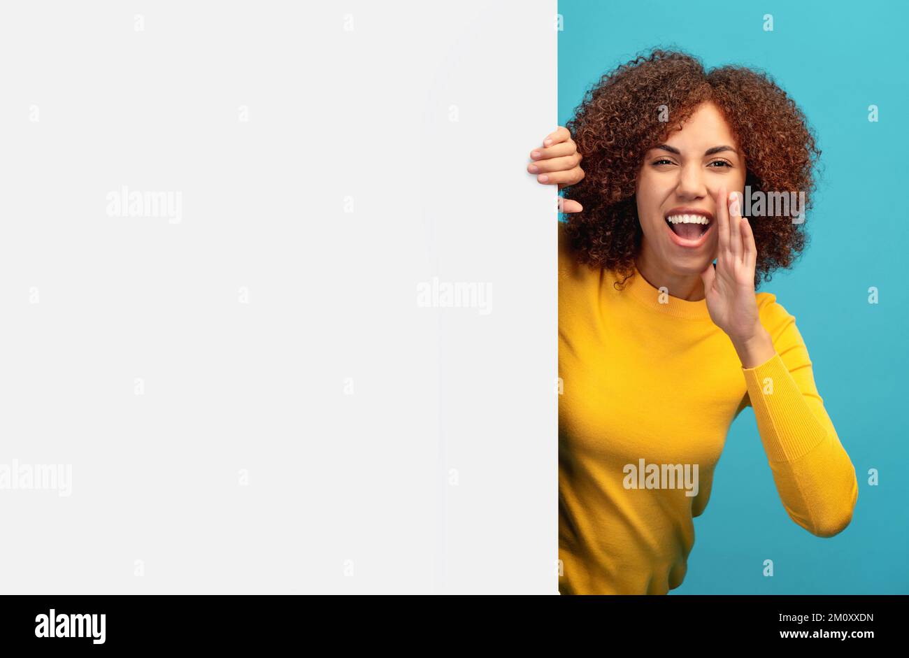 Young happy African American woman peeking out from empty white board and holding her hands near her mouth scream Stock Photo