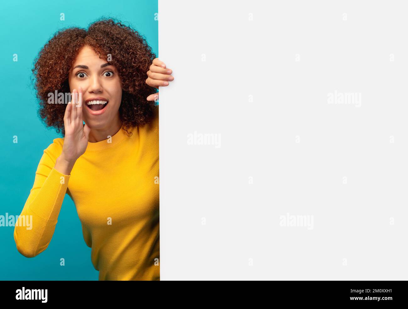 Young beautiful African American woman peeking out from empty white board and holding her hands near her mouth scream Stock Photo